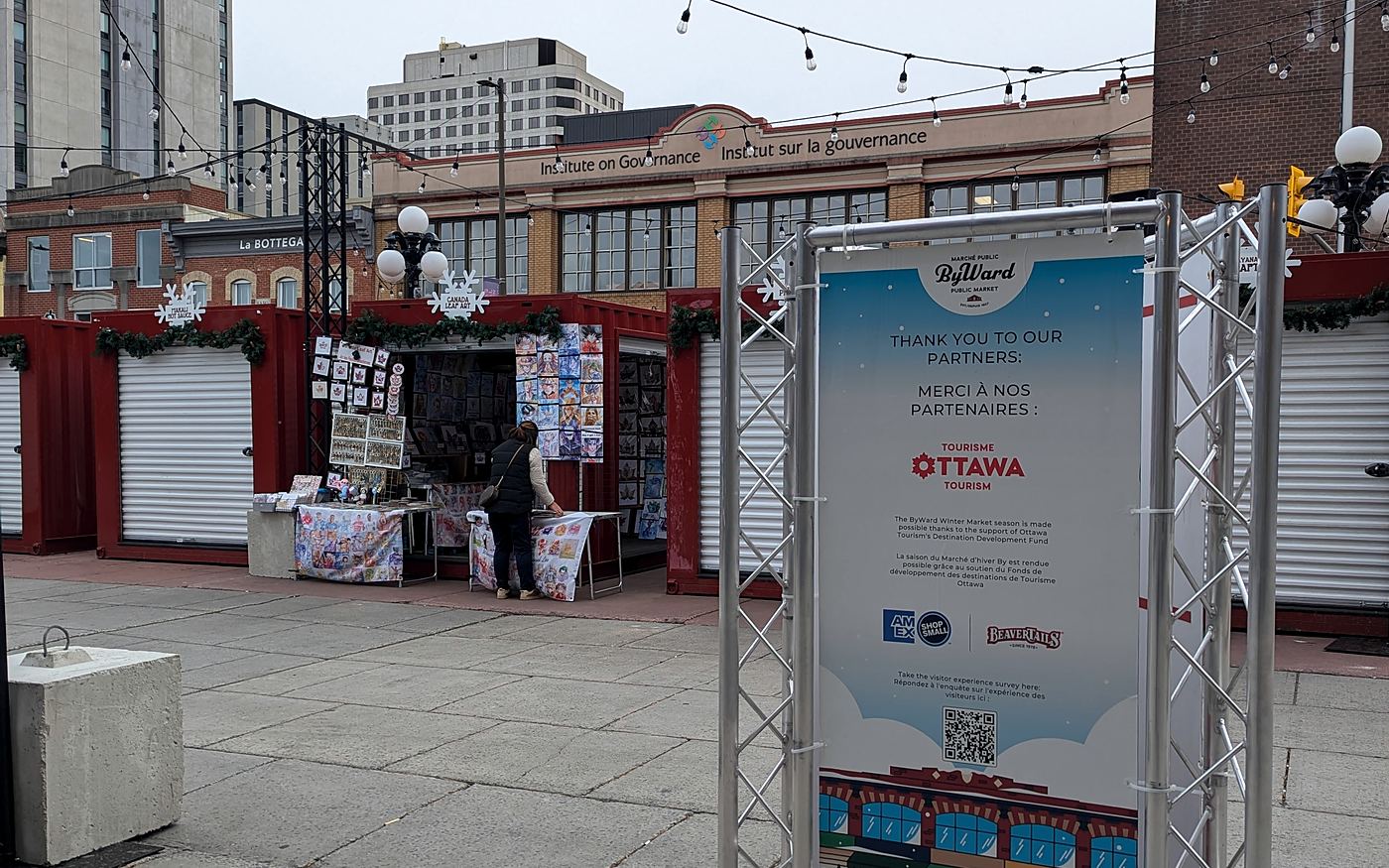 A ByWard Winter Market sign thanking Ottawa Tourism's Destination Development Fund, with market stalls behind