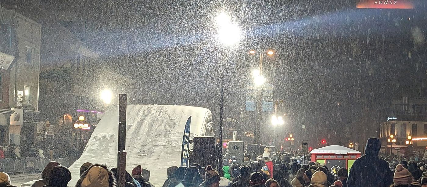 A crowd in falling snow at night facing the lit Barbegazi snowmobile ramp on a ByWard Market street during Winterlude